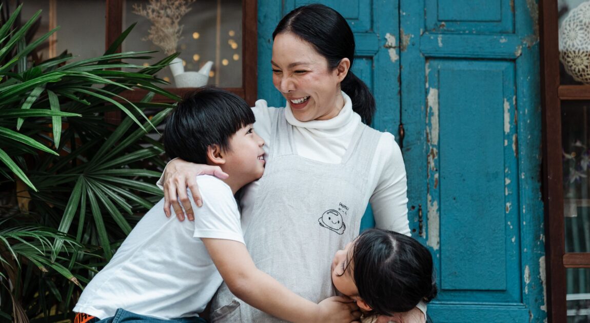 Happy cheerful mother bonding with cute son and daughter in casual wear while standing together against old shabby blue door and green garden plants
