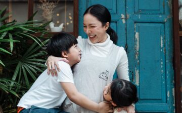 Happy cheerful mother bonding with cute son and daughter in casual wear while standing together against old shabby blue door and green garden plants